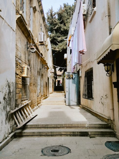 A residential street scene during a home relocation process showing a narrow street with parked cars lining both sides. In the center, a flatbed trolley loaded with large, wrapped cardboard boxes and